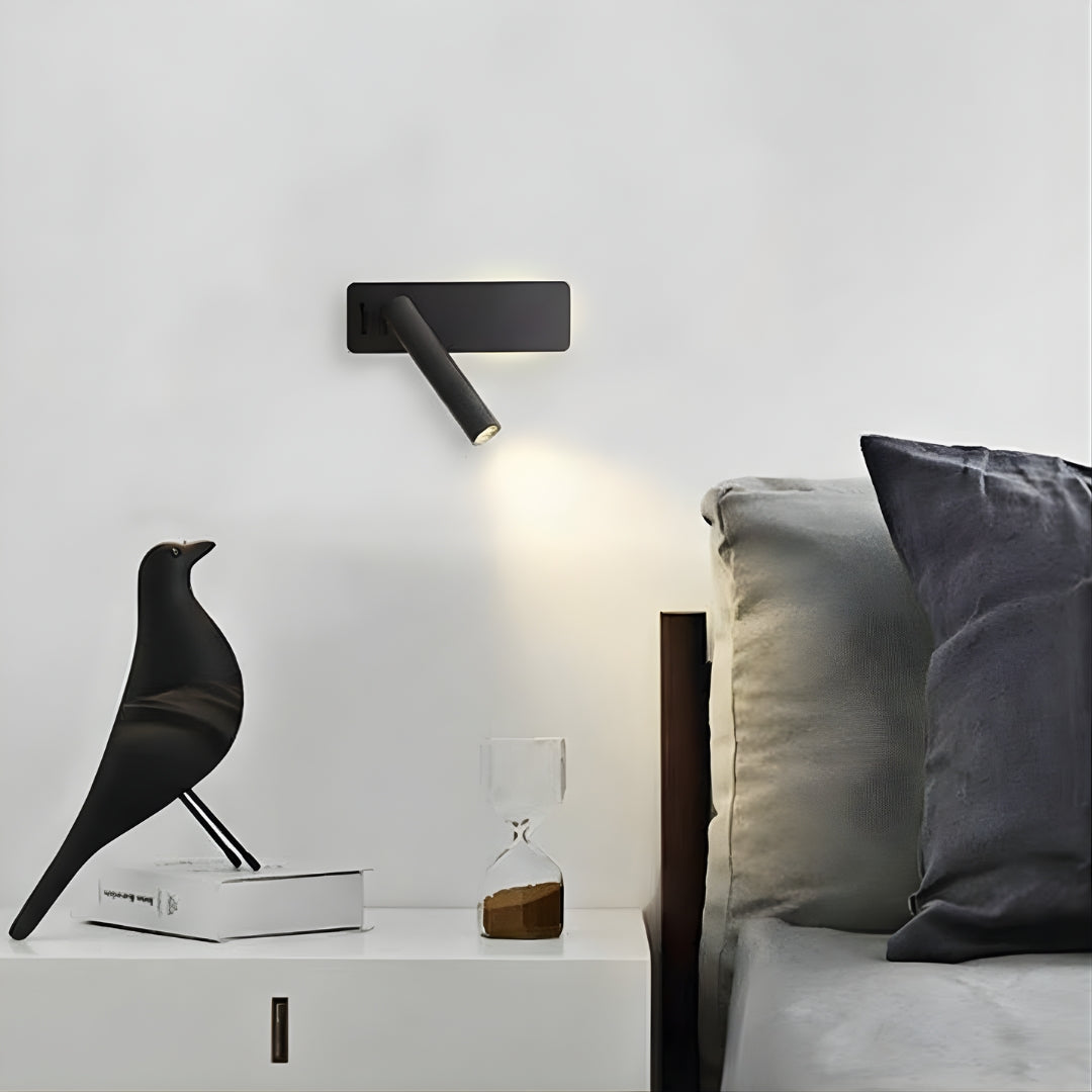 Czarna lampa ścienna na białej ścianie nad stolikiem nocnym z książką, czarną figurką ptaka i klepsydrą.