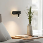 Czarna lampa ścienna nad brązowym stolikiem nocnym z książką, telefonem i rośliną.
